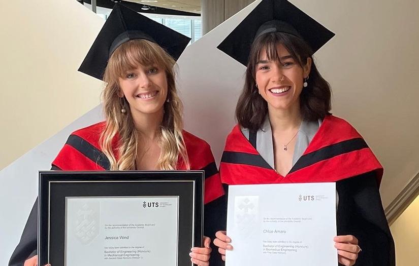 Two smiling UTS graduates holding up their qualifications, dressed in the graduation attire.