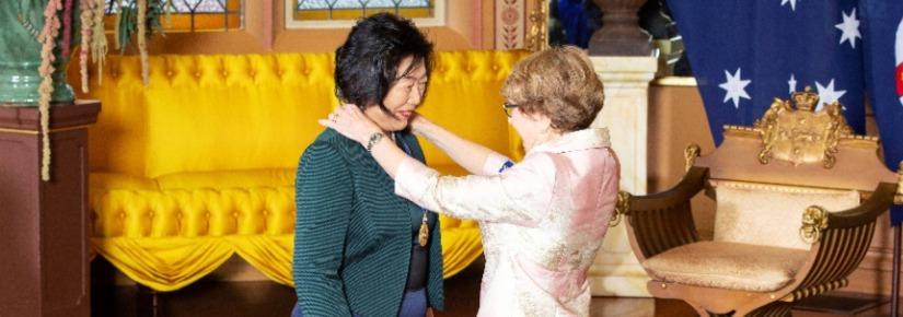 Jie Lu receiving her Order of Australia medal from the NSW Governor General, Margaret Beazley. The Governor General is placing the medal around Jie Lu's neck. Jie Lu is smiling, the Governor General's side profile is visible and she also appears to be smiling. Jie Lu is wearing a green fitted jacket on top of a black, navy and green striped dress. The Governor General is wearing a light pink satin fitted dress suited,