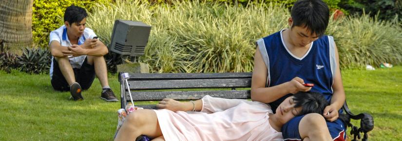 A woman lounges on a man's lap as he looks at his iPhone on a park bench.