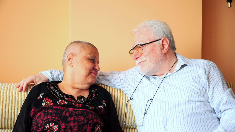 Lady and her husband sitting on a couch side by side smiling at each other. The lad is wearing a long sleeved black top with a red pattern on the front. She has lost her hair due to cancer treatment. The main is wearing a striped button up shirt. He has grey hair and glasses. 