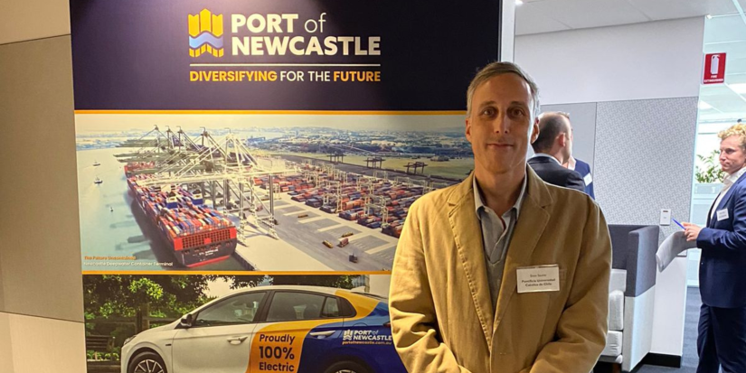 Professor Enzo Sauma stands against pull up banner promoting the Port of Newcastle. The banner features a brightly lit photo of a dock and the sea.