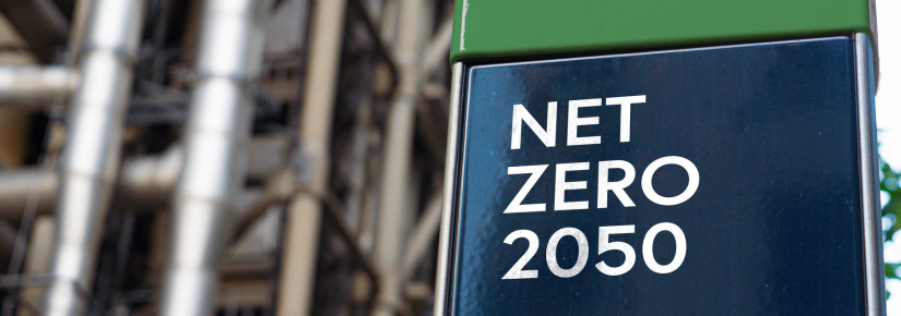 A blue and green shiny block says "NET ZERO 2050" in white capital letters. Pipes and infrastructure can be seen blurry in the background.