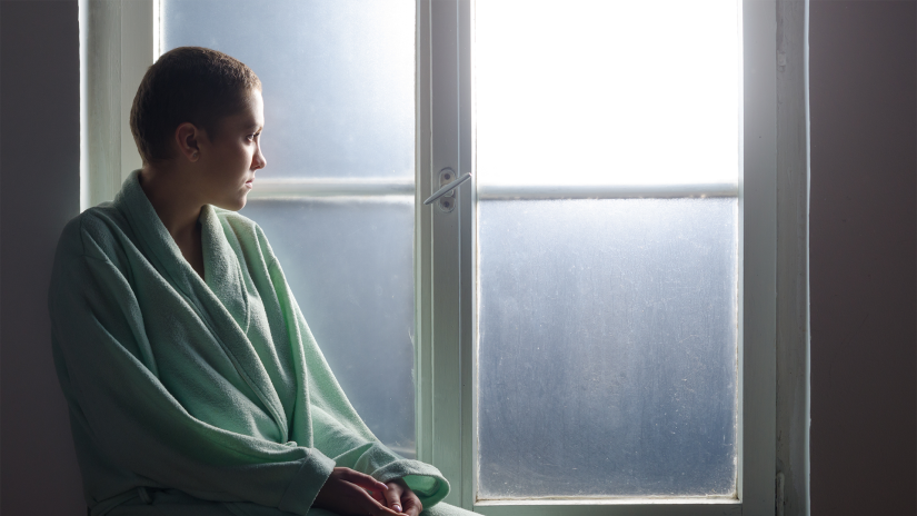 Young woman with short hair sits in front of window. She is wearing a bath robe and looking out the window, which has frosted glass. 