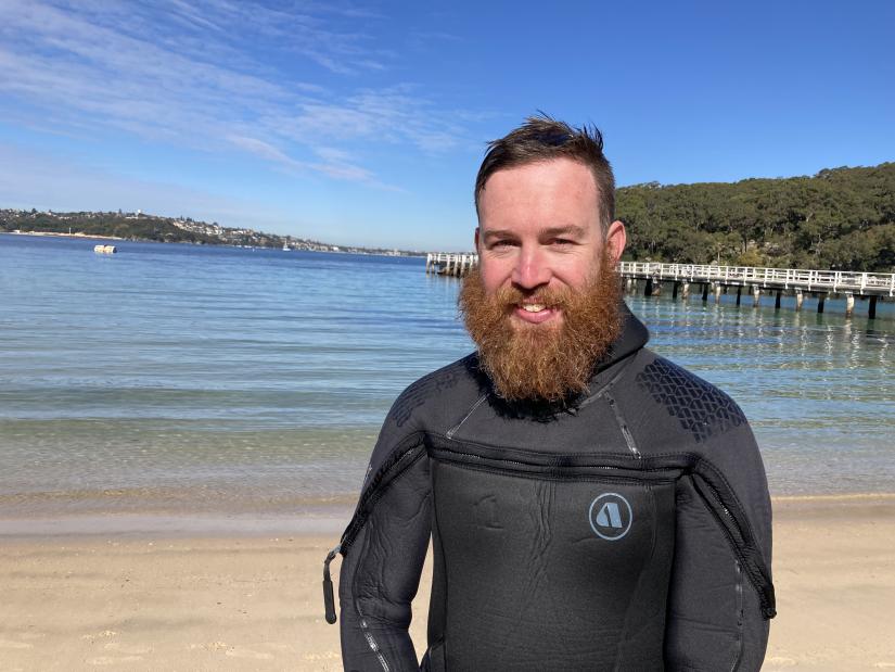 A man in a wetsuit stands on the beach with the ocean behind him