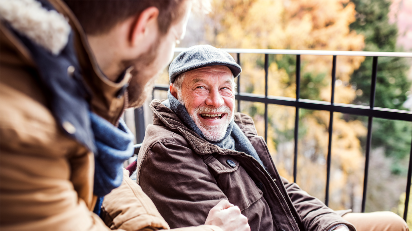Father and son, both with beards, on a walk. The father is wearing a flat cap, scarf and coat. The son is wearing a scarf and coat. The are looking at each other and smiling. 