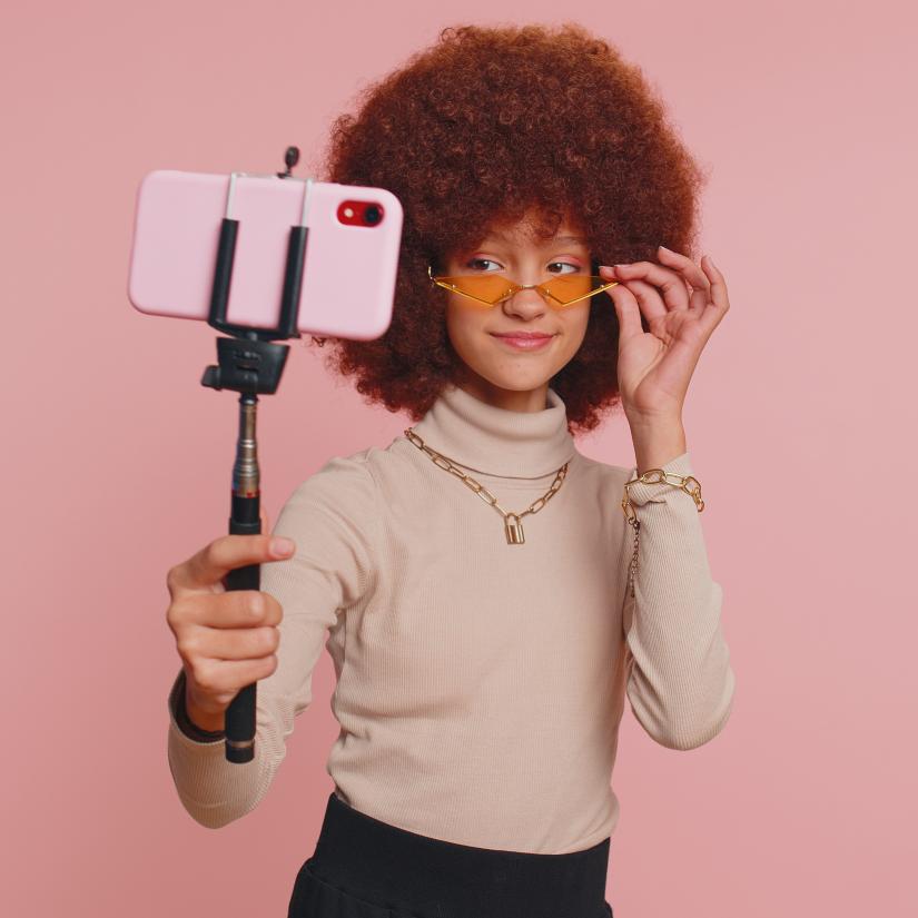 A young woman holds a camera held on a selfie stick up to face her 