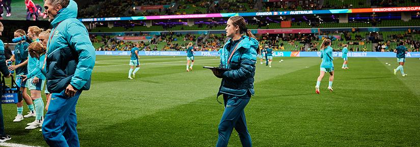 A woman dressed in a teal uniform holds a clipboard on the sidelines of a football pitch, players training in the background.