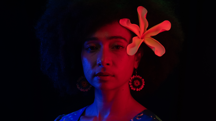 A woman with a flower in her hair looks at the camera bathed in red and blue lights
