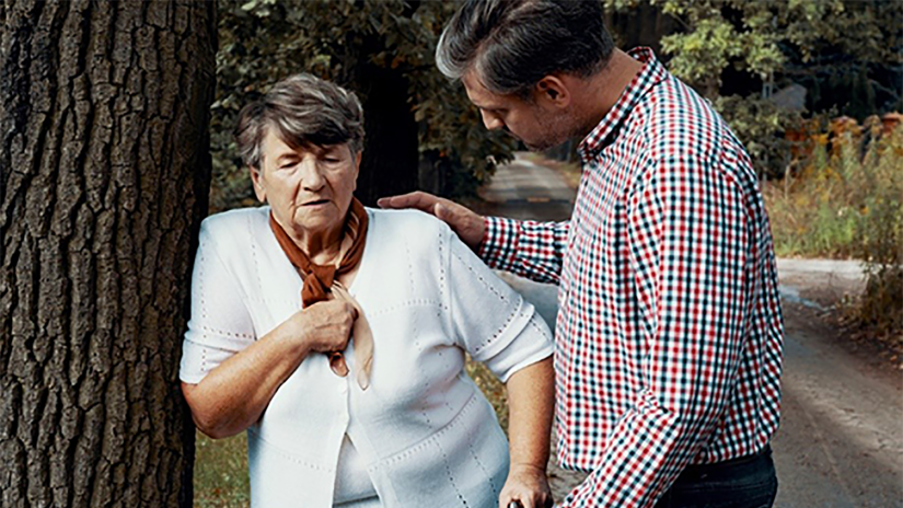 Older lady is leaning on a tree with he hand on her chest. A middle aged man has his hand on her shoulder to comfort her. 