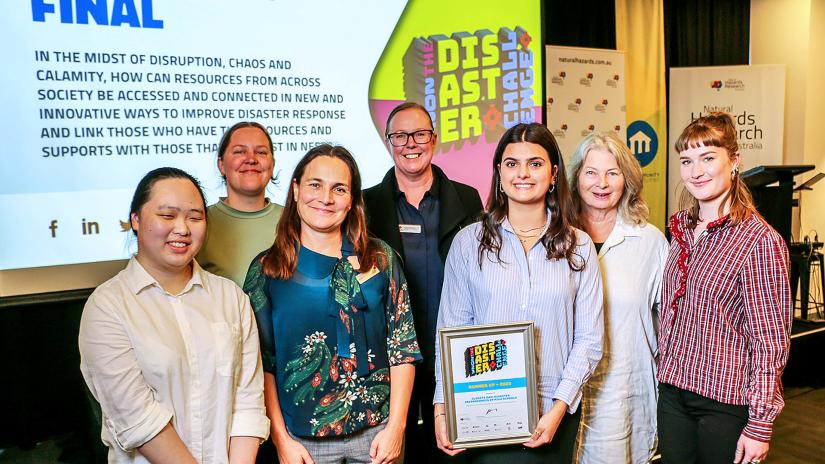 6 people of mixed genders and ages stand in front of a screen. A female UTS students holds the award won by her team for the Disaster Challenge.