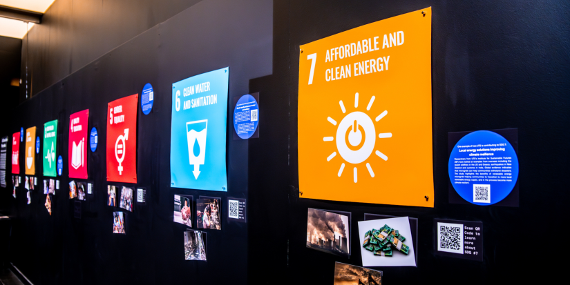 In the right half of the image, against a black wall a square orange sign reads “7 Affordable and Clean Energy” with an ‘ON’ button symbol. There are more colourful signs further to the left, all with various photos pinned beneath each of them.