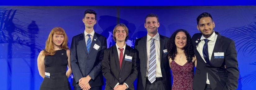 Six students - four male, two female - stand against a rich blue backdrop. They are wearing black suits and dresses, smiling. From left to right: Madeleine McWlliam; Cooper Crellin; Callum Burke; Matthew Dutaillis; Samantha Morris; and Sai Muthukumar.