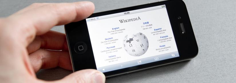 A person holds a mobile phone which displays the Wikipedia homepage.