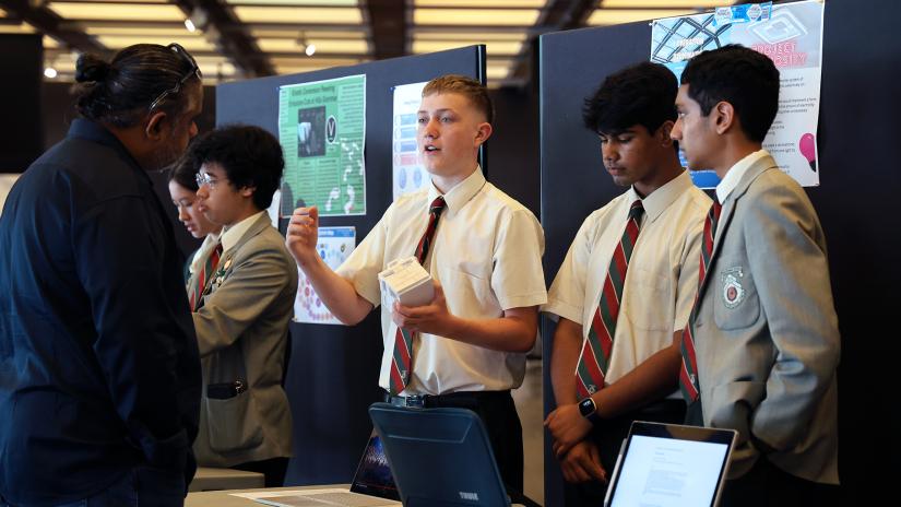 Several Year 10 students (all genders) stand behind a desk while showcasing their presentations to other students and teachers