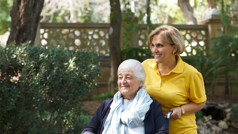 Caregiver with blonde hair and a yellow polo shirt is pushing an older lady with grey hair in a wheelchair. The lady in the wheelchair is wearing a navy jacket and a paly blue scarf. They are both smiling.