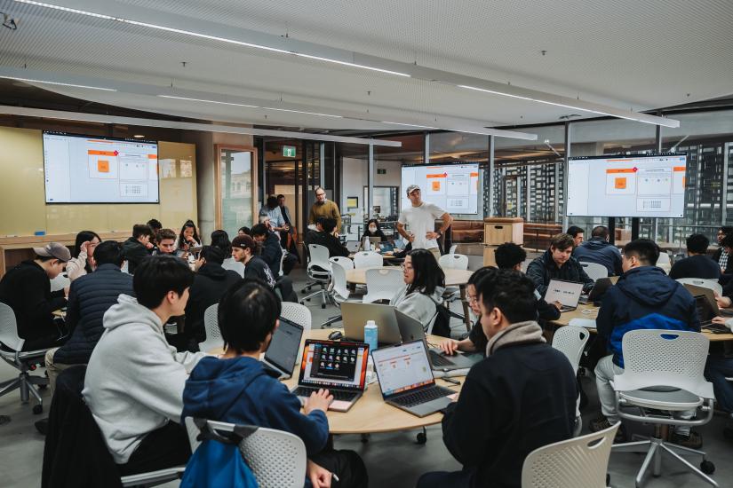 Image of students participating in the iOS Hackathon. Students are working in groups at round tables in the Apple Foundation Room in Building 11.