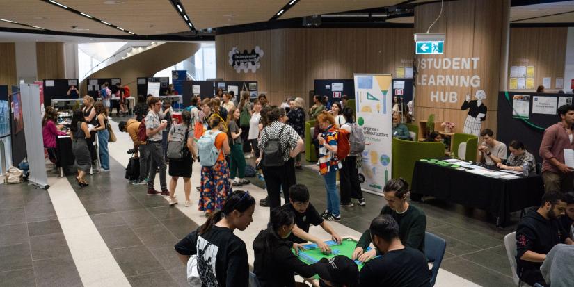 A wide shot of the Student Learning Hub in UTS Building 2 shows students wandering the 2024 Think Global Fair. Students can be seen playing Mahjong in the foreground. A cluster of grey booths can be seen in the background.