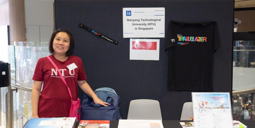 Hsiao Ching (left) poses in a deep red t-shirt with letters "NTU" printed. The backdrop features a sign that reads "Nanyang Technological University (NTU) in Singapore". An image of the Singaporean flag features below it.