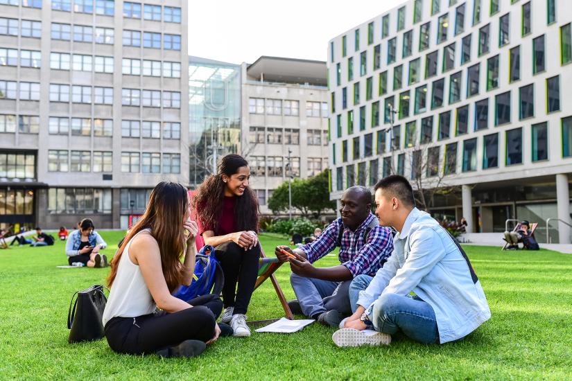 a group of students sit on the lawn, university buildings in the backgrund