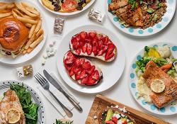 Plates of brightly coloured food cover a table, with means including fish, salads, pizza, a burger and chips, and strawberry and ricotta on toast.
