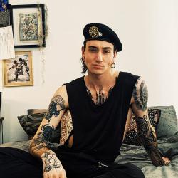 Jason Clark. Casual pose facing camera in his bedroom. Black beret, sleeveless black top, tattoos. 