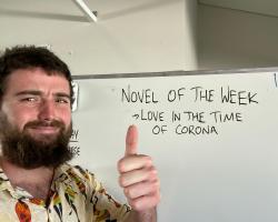 A young man stands in front of a white board smiling and holding his thumb up. The white board has written on it "Novel of the week: Love in the time of Corona"