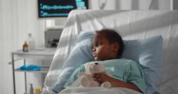Sick African-American girl wearing blue hospital gown lying in hospital bed with white sheets and hugging teddy bear