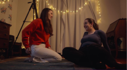 two young woman sitting on lounge room floor talking and smiling. There's video camera equipment behind them.