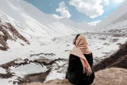 A woman in a headscarf sitting on a rock in an alpine environment looking at the snow covered landscape in front of them