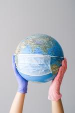 Image of a globe with a disposable mask on it being held between two hands one with a pink glove and one with a blue glove. 