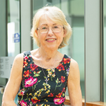 Imelda Gilmore has short blonde hair and is wearing sleeveless top that is black with a flora print. She wears glasses and is smiling at the camera.