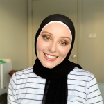 Nadine El-Kabbout is smiling at the camera. She is wearing a white top with black horizontal stripees and a black head scarf.