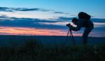 A male with backback leaning down towards camera on tripod with a background of dark sunset and clouds