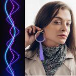 Image of a trans woman holding a purple earring to their ear. A purple voice sound wave is to the left of them.